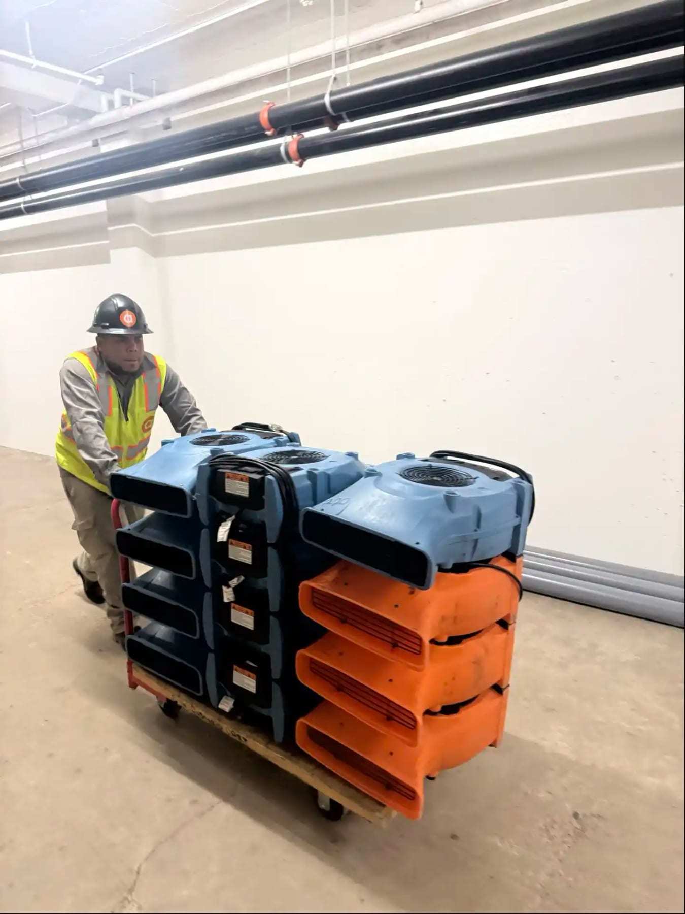 Worker in a hard hat pushing a cart stacked with air movers across a garage