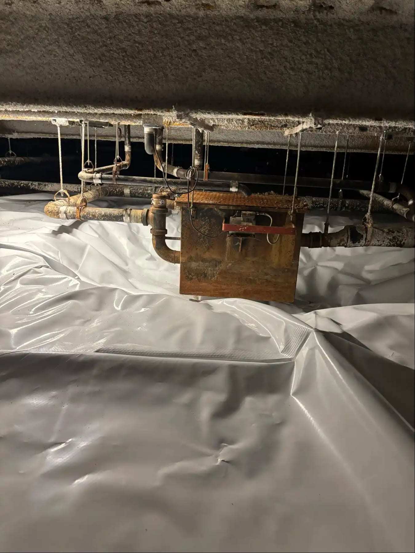 Crawlspace with a vapor barrier below rusted pipes and a hanging box