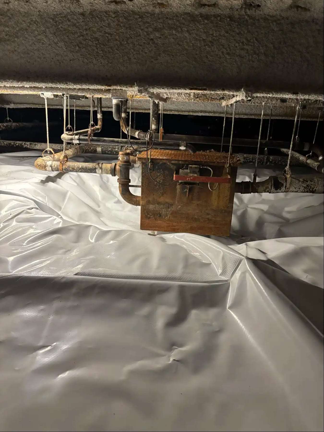 Crawlspace with a vapor barrier below rusted pipes and a hanging box