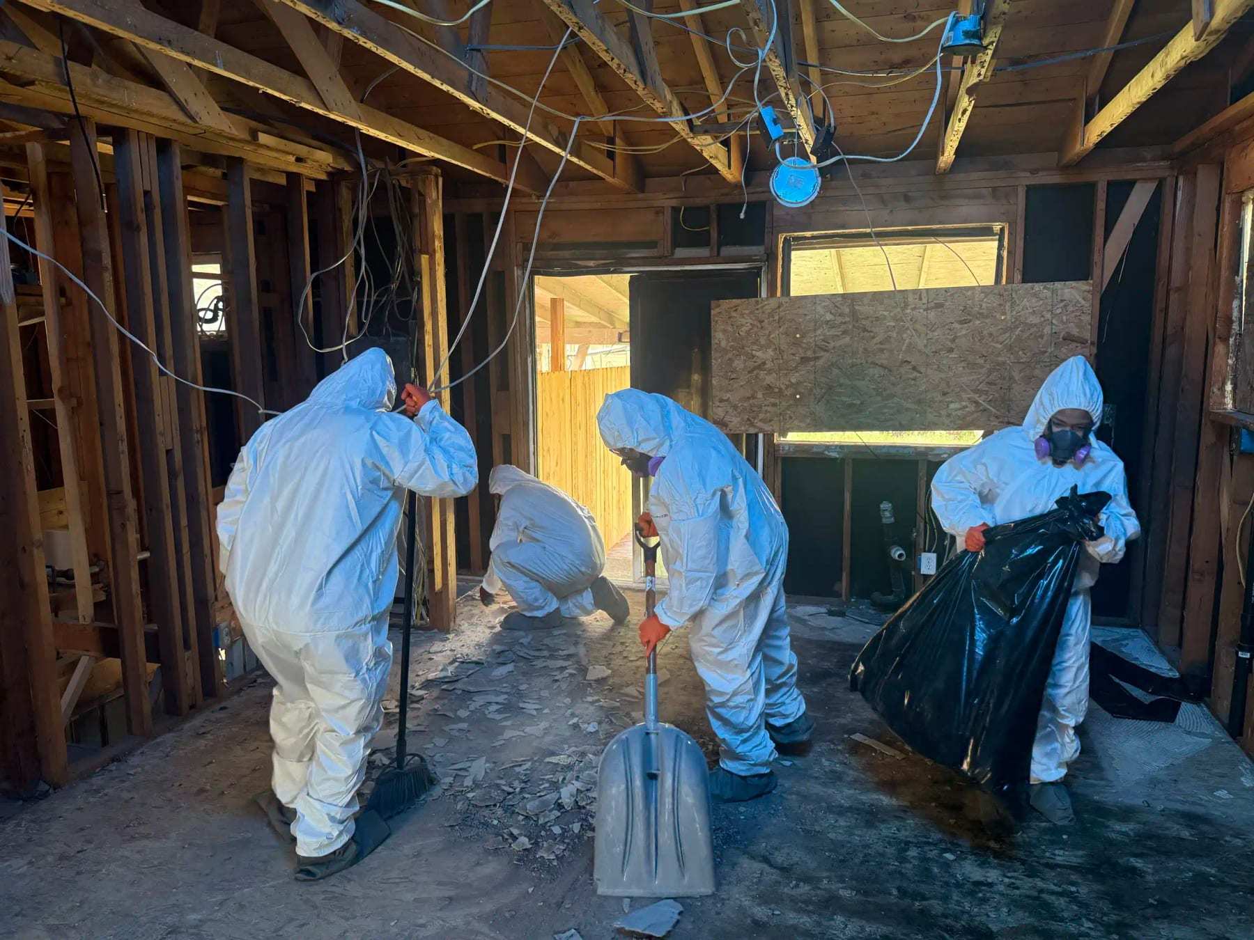 Four technicians in protective suits shoveling debris in a basement stripped to the framing