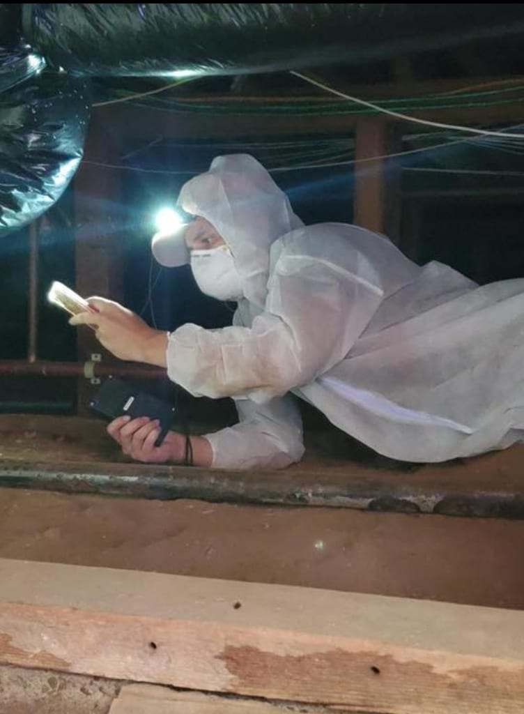 Technician in a protective suit inspecting a crawlspace with a flashlight