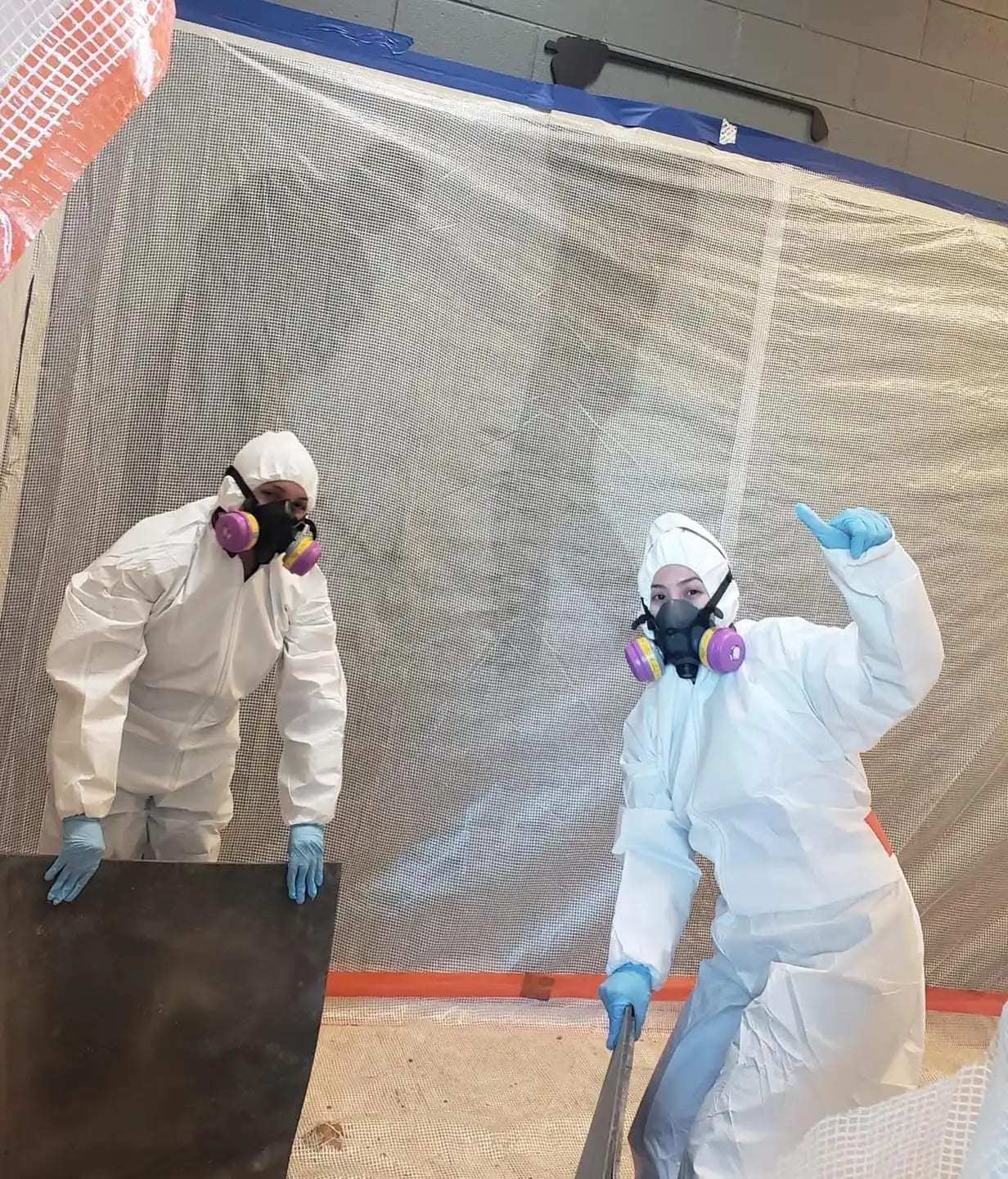 Two technicians in protective suits and respirators at a plastic containment wall